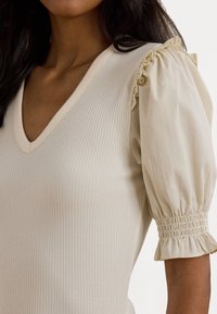 Kvinna iklädd en krämfärgad ribbad V-ringad topp med puffärmar som är samlade vid axlarna och ärmsluten.