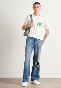 Camiseta de algodón blanca con el logo 'CK' en verde, combinada con jeans azules desgastados con rasgones en las rodillas y zapatillas blancas. Incluida una bolsa de hombro gris.