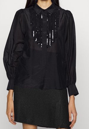 Kvinde iført en sort gennemsigtig bluse med lange ærmer med lodrette sorte palietter foran, kombineret med et tekstureret sort strikket skørt.