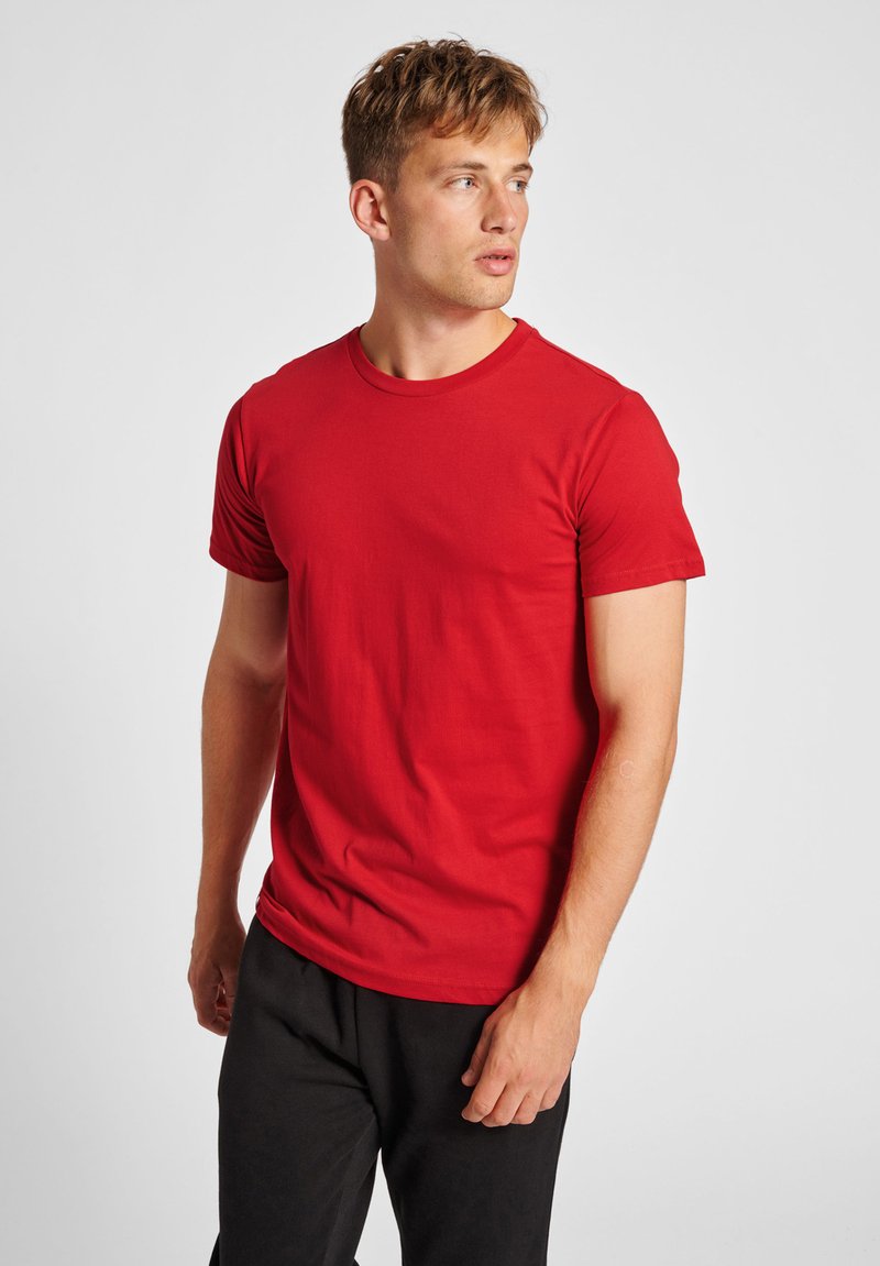 Rød bomulds-t-shirt med korte ærmer og rund hals. Glat tekstur, tætsiddende facon, ingen synlige mønstre, parret med sorte bukser.