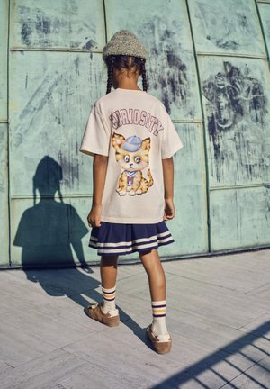 Bambino con capelli intrecciati che indossa una maglietta beige "CURIOSITY" con grafica di gatto, gonna blu scuro, calzini a righe e cappello, in piedi davanti a un muro di metallo usurato.