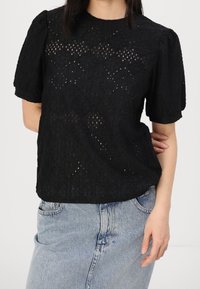 Top in pizzo nero con maniche a sbuffo corte, caratterizzato da intagli a motivo diamante e scollatura rotonda, abbinato a jeans in denim azzurro chiaro.