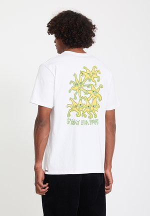 Man met krullend haar die wegkijkt, draagt een wit shirt met zes gele zeesterrenillustraties en groene tekst "Fishy Star Party" op de achterkant.