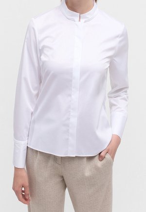 Donna che indossa una camicia bianca a maniche lunghe con collo alla coreana e pantaloni beige lavorati, una mano in tasca.