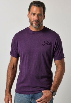 Uomo con capelli corti e scuri e barba, indossa una maglietta viola con maniche arrotolate e jeans, in piedi con una mano in tasca davanti a uno sfondo semplice.