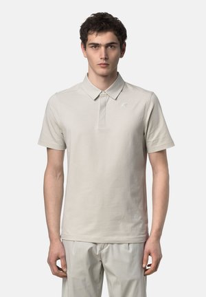 Giovane uomo in piedi che indossa una polo beige a maniche corte e pantaloni abbinati su uno sfondo chiaro uniforme.