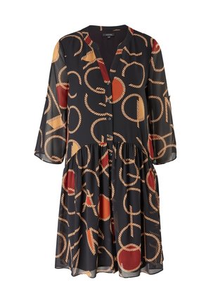 Mujer que lleva un vestido negro con grandes círculos rojos y beige y patrones de cuerda, combinado con botas de ante marrón hasta la mitad de la pantorrilla.