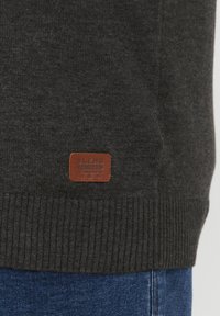 Pull en tricot gris foncé avec des poignets et un ourlet rayés. Présente une patch en cuir marron avec la marque "BLEND" sur la partie inférieure.