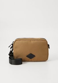 Tan rectangular crossbody bag with black adjustable strap and black logo patch on front, set against a plain white background.