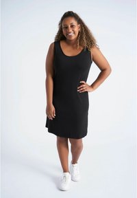 Robe noire sans manches avec un décolleté arrondi, confectionnée dans un tissu lisse, tombant juste au-dessus du genou, associée à des baskets blanches.
