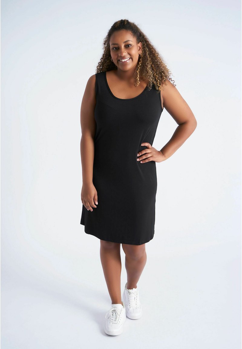 Robe noire sans manches avec un décolleté arrondi, confectionnée dans un tissu lisse, tombant juste au-dessus du genou, associée à des baskets blanches.