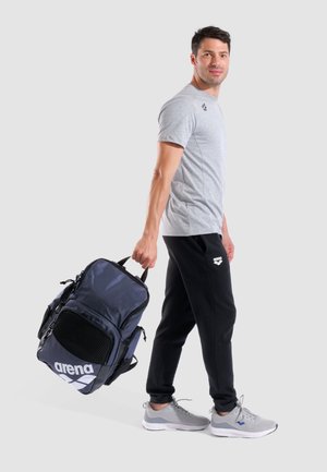 Zaino sportivo blu marina con accenti in rete e logo bianco, portato da una persona in t-shirt grigia e pantaloni neri, con scarpe da ginnastica grigie.