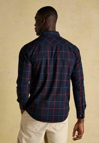 Koszula w kratę w odcieniach granatu z czerwonymi i niebieskimi wzorami, z zapięciem na guziki, kołnierzykiem typu button-down i długimi rękawami. Teksturowana tkanina; luźny krój. Jasne spodnie.