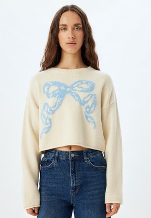 Suéter corto de crema con diseño de lazo azul, tejido texturizado, mangas anchas y escote redondo, combinado con jeans de mezclilla de cintura alta.