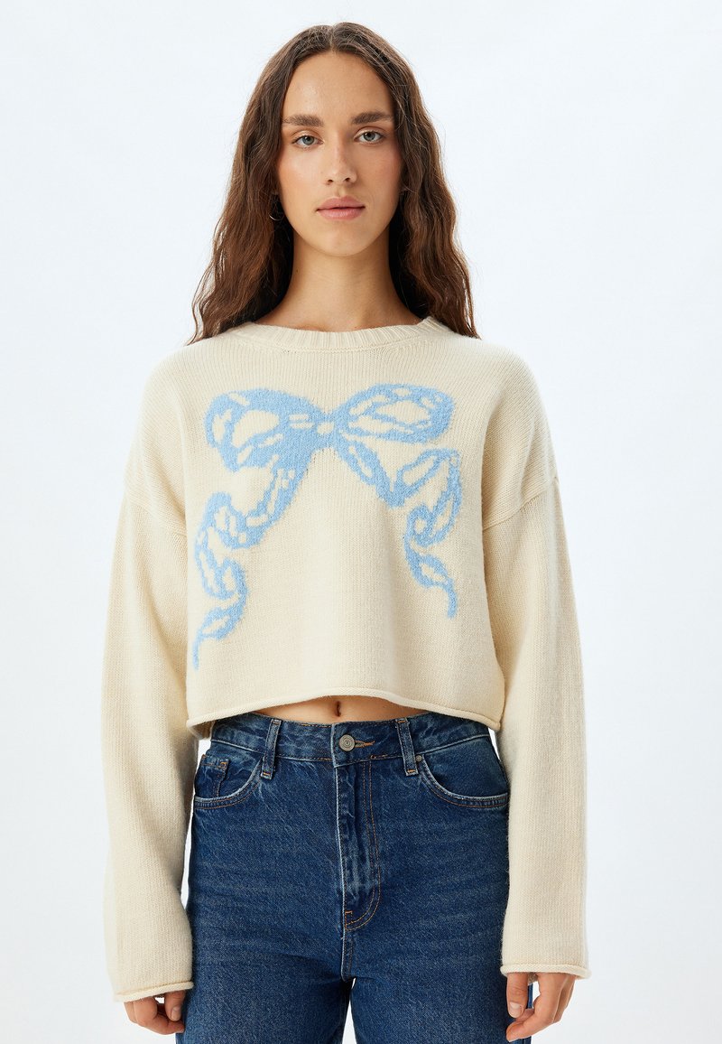 Maglione corto crema con motivo a fiocco blu, lavorazione a maglia testurizzata, maniche ampie e scollatura rotonda, abbinato a jeans in denim a vita alta.