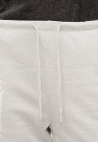 Κοντινή λήψη μέσης ανοιχτόγριζου παντελονιού φόρμας με λευκά κορδόνια δεμένα πάνω σε σκούρο γκρι τοπ.