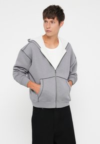 Pelēka kapuču džemperis ar baltu iekšējo kapuci, tajā ir divas priekšējās kabatas un kontrastējoša šuve gar šuvēm. Mīksts kokvilnas materiāls.