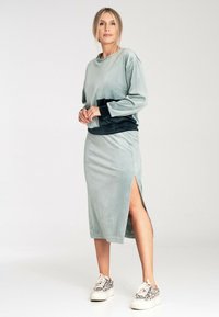 Jasnozielony welurowy sweter z czarną talią, w połączeniu z pasującą spódnicą z bocznym rozcięciem, noszony z wzorzystymi sneakersami.