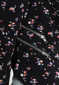 Tejido negro con textura floral y pequeñas flores en tonos rosa, morado y blanco; cuenta con dos bolsillos con cremallera plateada en una manga.