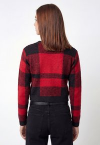 Red and black checkered sweater with a cropped design, knitted texture, and ribbed cuffs, worn with black jeans. Back view shown.