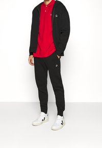 Czarna bluza z zamkiem na całej długości z regulowanym kapturem, czerwona koszulka polo, czarne dresy i białe sneakersy z czarnymi akcentami. Posiada naszyte logo.