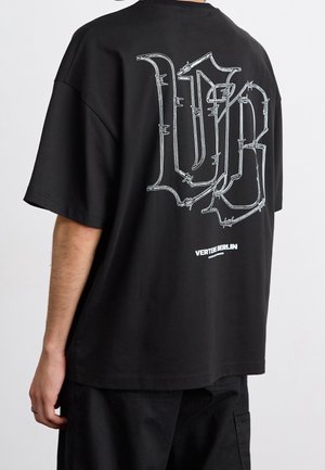 Osoba nosząca czarny oversize T-shirt z dużymi białymi literami graffiti w stylu drutu kolczastego na plecach oraz małym napisem poniżej o treści Verte Berlin.