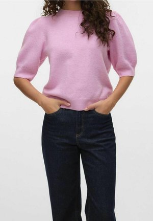 Frau trägt einen hellrosa kurzen Pullover mit Puffärmeln und dunkelblaue hoch taillierte Jeans, Hände in den Taschen.