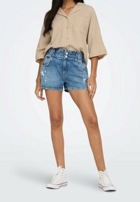 Shorts en denim bleu moyen avec une taille haute, des détails effilochés et des ourlets retroussés, associés à une chemise beige texturée et des baskets blanches.