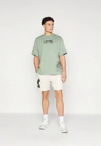 Πράσινο βαμβακερό μπλουζάκι με μαύρα γραφικά patches, συνδυασμένο με κρεμ σορτς που έχουν μαύρο γραφικό. Το μοντέλο φοράει λευκά αθλητικά παπούτσια.