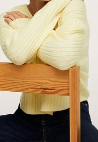 Maglione a coste giallo chiaro con maniche lunghe, realizzato in morbido tessuto a maglia, indossato incrociato sulle braccia. Sedia di legno visibile in primo piano.