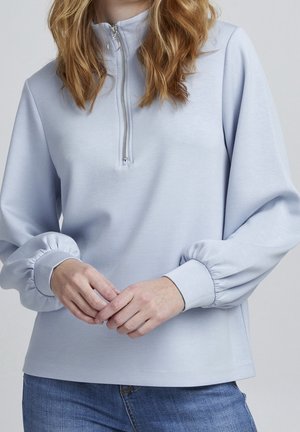 Femme portant un pull bleu clair à manches longues avec une demi-fermeture éclair et des poignets élastiques, associé à un jean bleu, mains légèrement jointes.