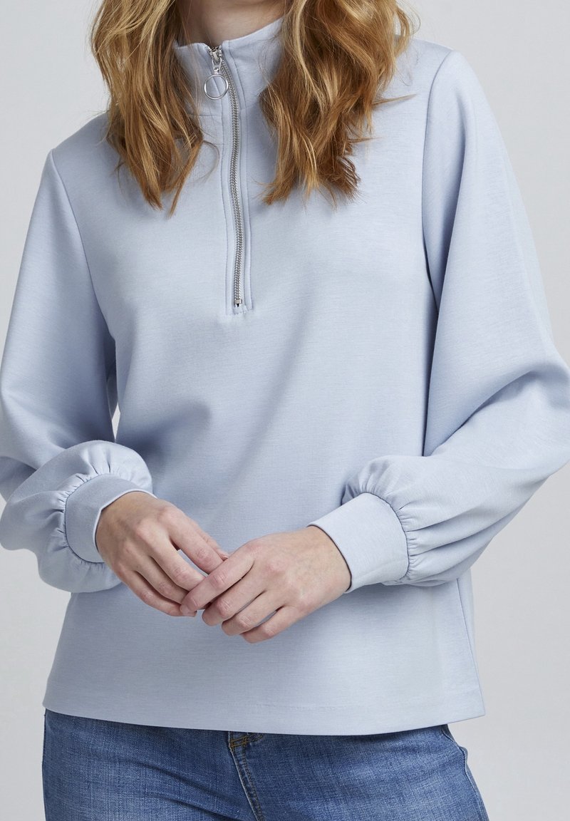 Femme portant un pull bleu clair à manches longues avec une demi-fermeture éclair et des poignets élastiques, associé à un jean bleu, mains légèrement jointes.