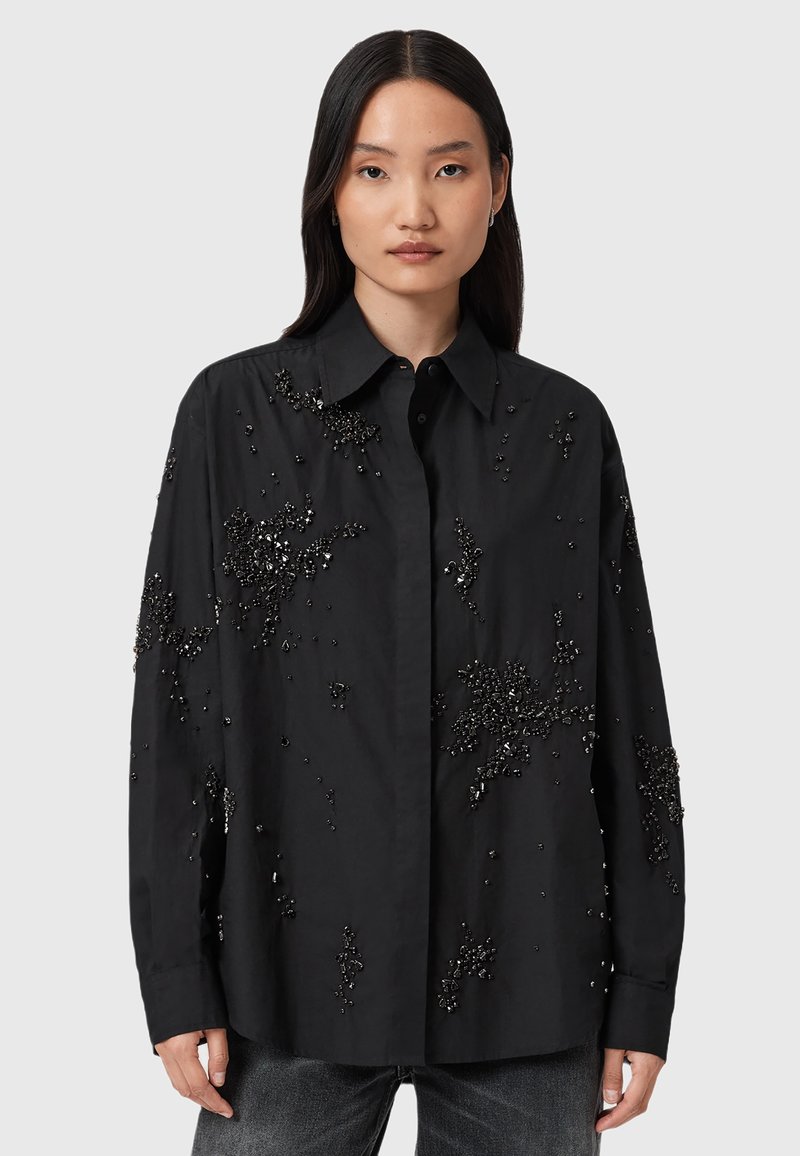 Camicia nera in tessuto con una texture liscia, caratterizzata da decorazioni nere sparse e un colletto con bottoni.