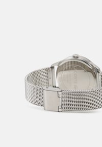 Pulsera de malla plateada con caja de acero inoxidable. Presenta una esfera redonda, bisel liso y logotipo de la marca grabado en el cierre.