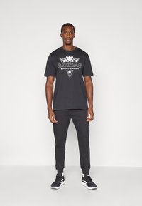 Juodas medvilninis marškinėliai su balta "ADIDAS SPORTSWEAR" logotipu ir grafiniu dizainu, derinami su juodomis kelnėmis ir juodomis sportinėmis bateliais.