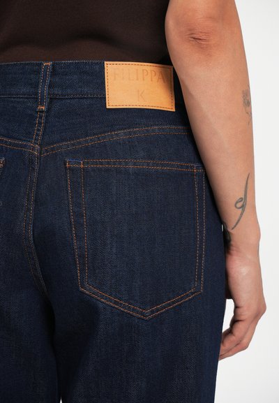 Jeans de mezclilla azul oscuro con un parche de cuero marrón claro etiquetado "FILIPPA K" en la cintura, presentando costuras naranjas y un clásico diseño de cinco bolsillos.