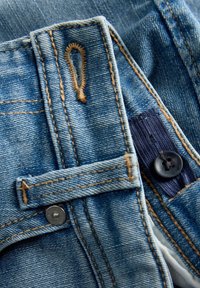 Gros plan sur un jean en denim bleu montrant un bouton, une boutonnière, une passants de ceinture et des détails de couture orange contrastante.