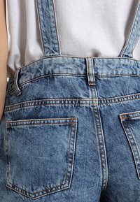 Bakvy av en person som bär ljusblå denimöveraller med två bakfickor och en vit skjorta instoppad.