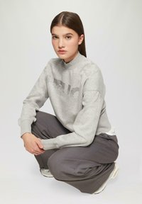 Grijze sweatshirt met een hoge hals en gemetste letters op de voorkant, gecombineerd met donkergrijze wijde broekspijpen en lichtgekleurde sneakers.