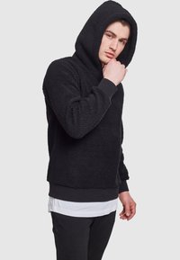 Felpa nera in pile con superficie strutturata, cappuccio e polsini a coste. Indossata sopra una camicia bianca a strati e abbinata a pantaloni neri.