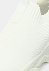 Λευκό πάνινο παπούτσι χωρίς κορδόνια με υφή στο πάνω μέρος, ριμπ λεπτομέρειες και μαλακή σόλα. Ο σχεδιασμός είναι μινιμαλιστικός με καθαρό φινίρισμα.