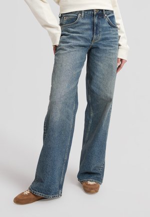 Person trägt weite blaue Jeans, weißes Langarmshirt und braune Sneakers vor einem schlichten hellen Hintergrund.