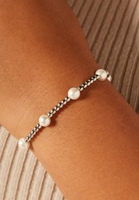 Armband met kralen van afwisselend witte parels en zilveren bolletjes, met een flexibel ontwerp en glanzende metalen accenten op een pols met huidskleur.