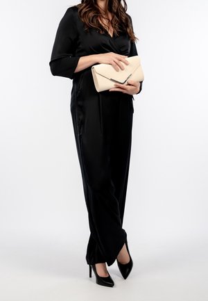 Mujer con mono de satén negro sosteniendo un bolso de mano beige, llevando zapatos de tacón altos puntiagudos negros, de pie frente a un fondo blanco liso.