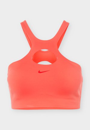 Koraalkleurige sportbeha met gekruiste schouderbandjes en een centrale uitsnijding onder de halslijn, met een klein rood Nike Swoosh-logo aan de voorkant.