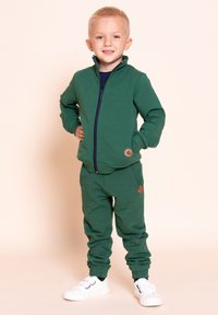 Młody chłopiec stoi z rękami w kieszeniach, ubrany w zieloną kurtkę na zamek, pasujące spodnie dresowe i białe trampki na beżowym tle.