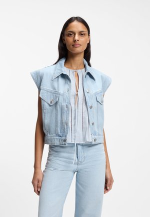 Donna che indossa un giubbotto di jeans senza maniche azzurro chiaro sopra una maglietta bianca, abbinato a jeans di jeans azzurro chiaro, in piedi contro uno sfondo bianco.