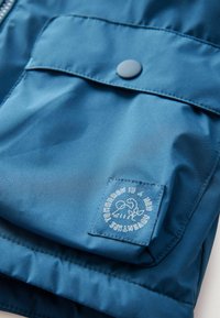 Blaue Nylonjacke mit einer Druckknopf-Tasche, gesteppten Akzenten und einem Logopatch mit Text. Glatte Textur mit matter Oberfläche.