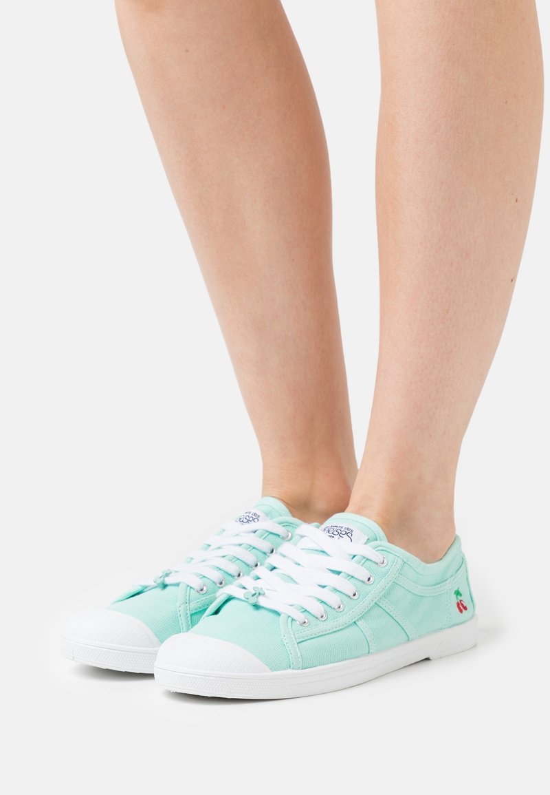 Mátově zelené plátěné tenisky s bílými gumovými podrážkami, vyznačující se bílými tkaničkami a malým vyšitým motivem třešně na boku.