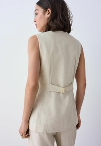 Beige ärmellose Blazer mit strukturiertem Finish, featuring ein Rückengurtdetail zum Raffen. Klare Linien und taillierte Passform für einen strukturierten Look.